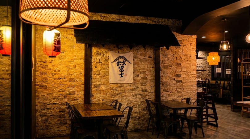 Warmly lit Japanese restaurant interior with stone walls, wooden tables, metal chairs, and hanging lanterns.